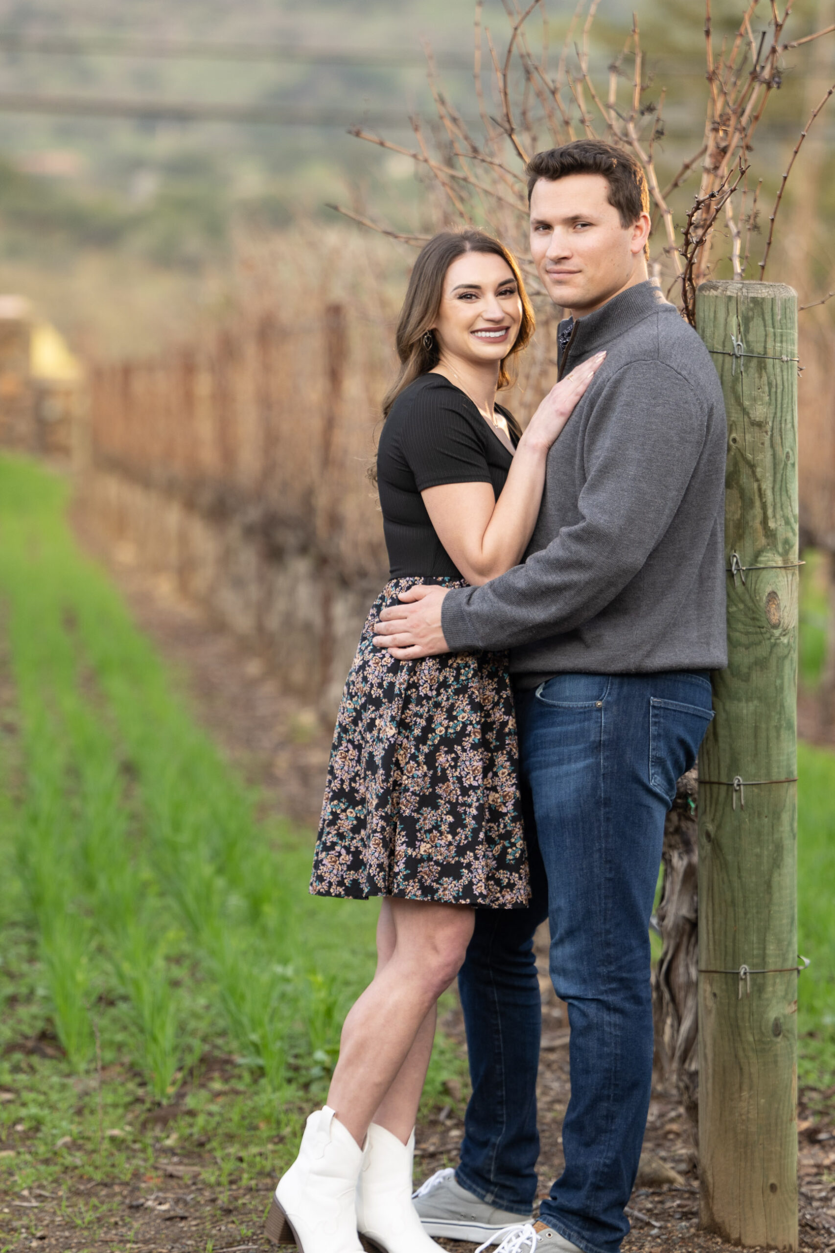A couple stands closely together in a vineyard. The woman wears a black top and floral skirt, while the man is dressed in a gray jacket and jeans. They are outdoors with rows of grapevines behind them.