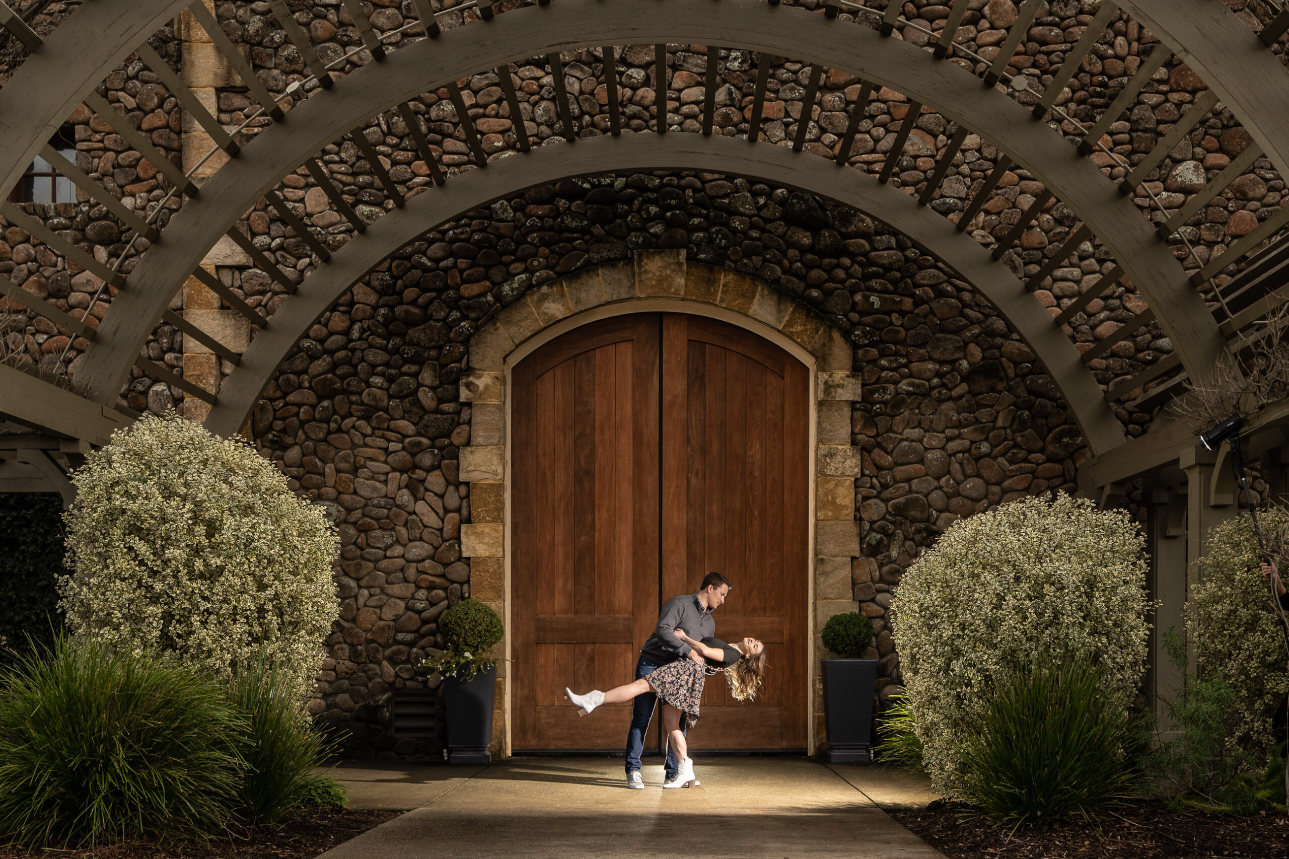 A couple poses in a dance dip under a large stone archway with wooden doors and greenery surrounding them.
