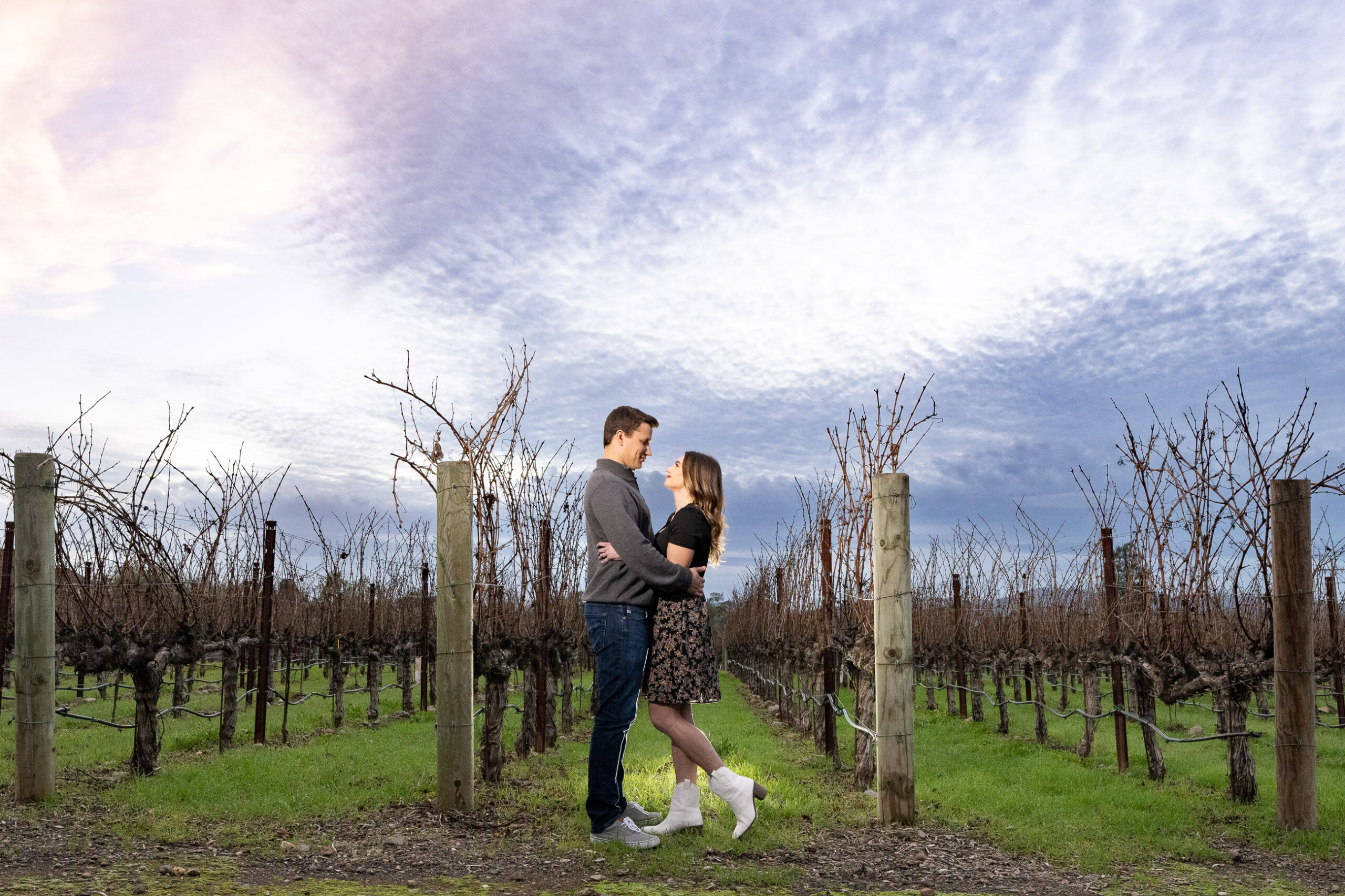A couple stands facing each other in a vineyard, with bare grapevines and an expansive sky with clouds in the background.