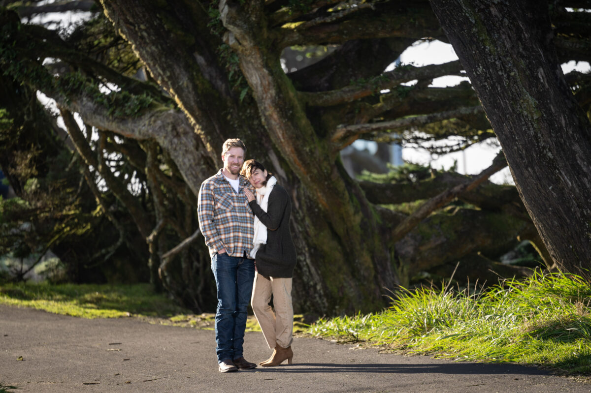 A man in a plaid shirt and a woman in a brown jacket stand close together on a path, with large, moss-covered trees in the background.