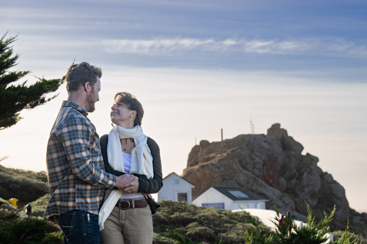 A couple stands close together, holding hands and smiling at each other with a rocky hill and small buildings in the background.