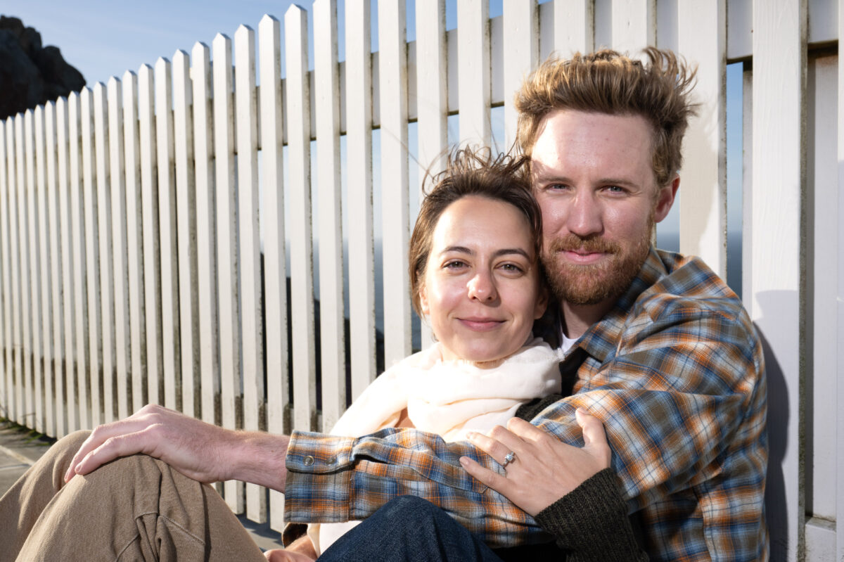 A man and woman sitting together, smiling, with the man's arm around the woman in front of a white picket fence.