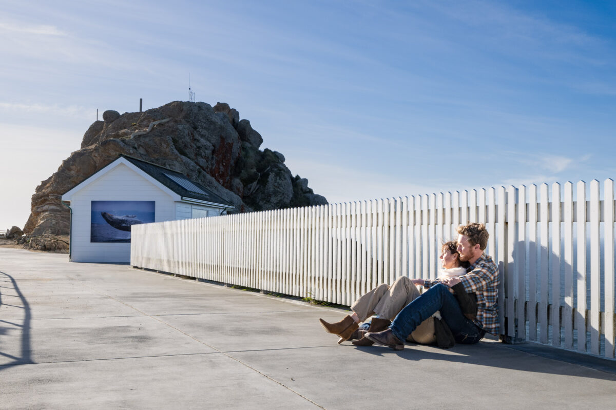 A couple sits on the ground, leaning against a white picket fence near a building with a whale mural, with a large rocky hill in the background on a clear day.