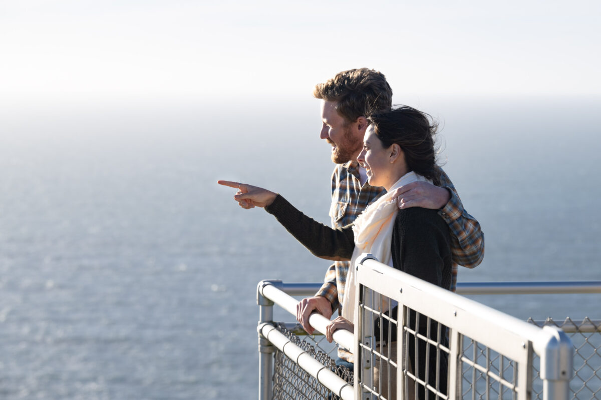 A couple stands on a metal balcony overlooking the ocean. The woman is pointing towards the horizon while the man has his arm around her.