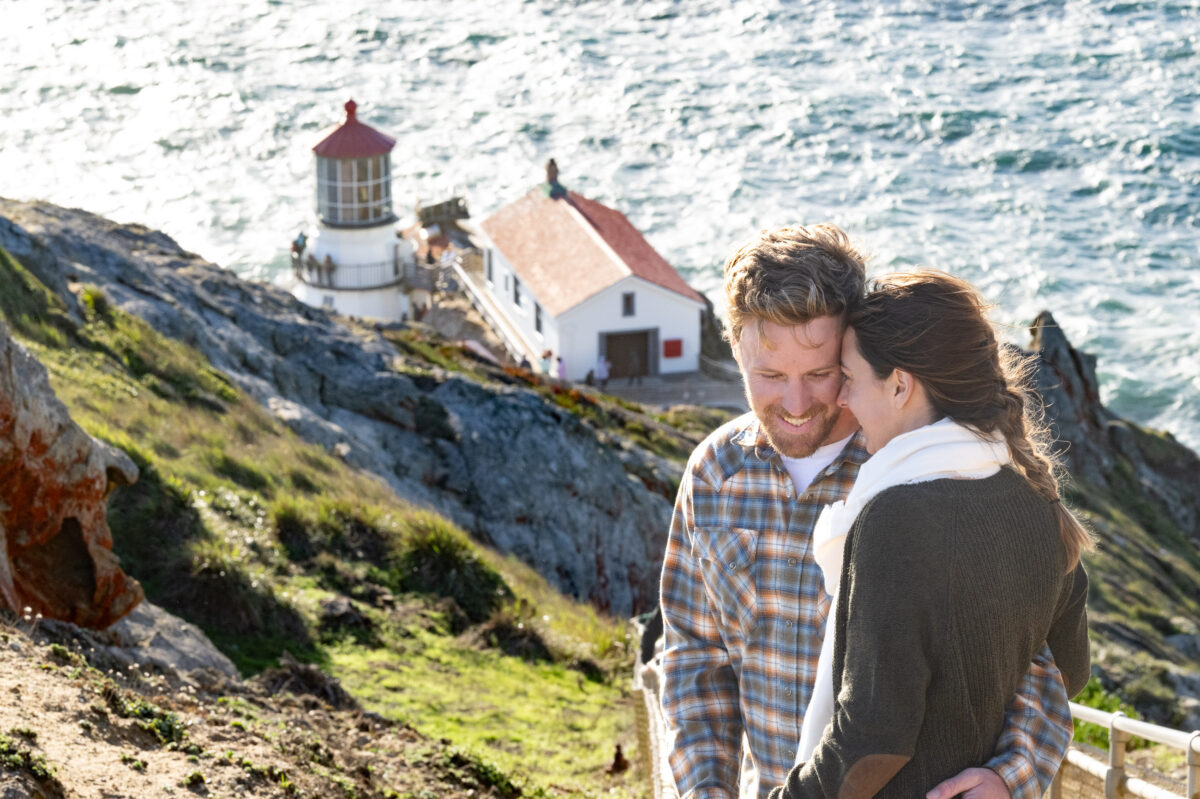 A couple stands close together on a rocky, grassy coastline with a lighthouse and the ocean in the background.