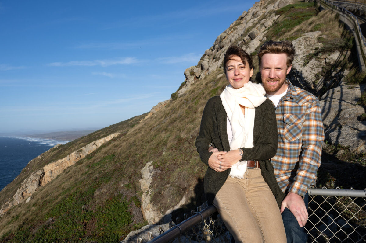 A woman and man smile while posing on a fence in front of a coastal cliff. The ocean is visible in the background under a clear blue sky.