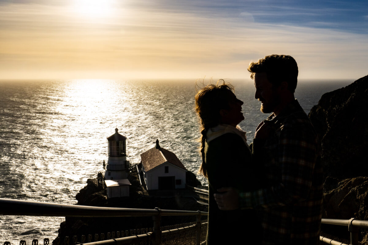 Two people stand close together, silhouetted against a sunset over the ocean, with a distant lighthouse and building visible on a cliff.