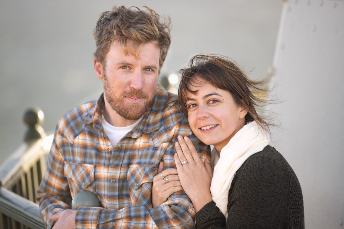 A man with a beard and plaid shirt stands beside a woman in a dark sweater and white scarf, both smiling and looking at the camera. The woman has her hand on the man's shoulder.