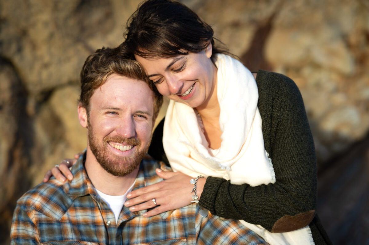 A man and a woman, both smiling, pose together in an outdoor setting. The woman has her hand on the man's shoulder, and they appear relaxed and happy.