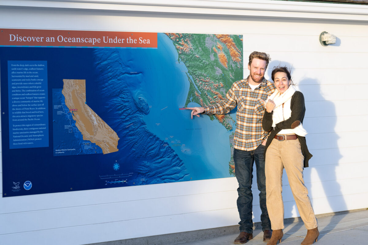 Two people stand next to an informational map display about the ocean, with the man pointing at a specific location on the map.