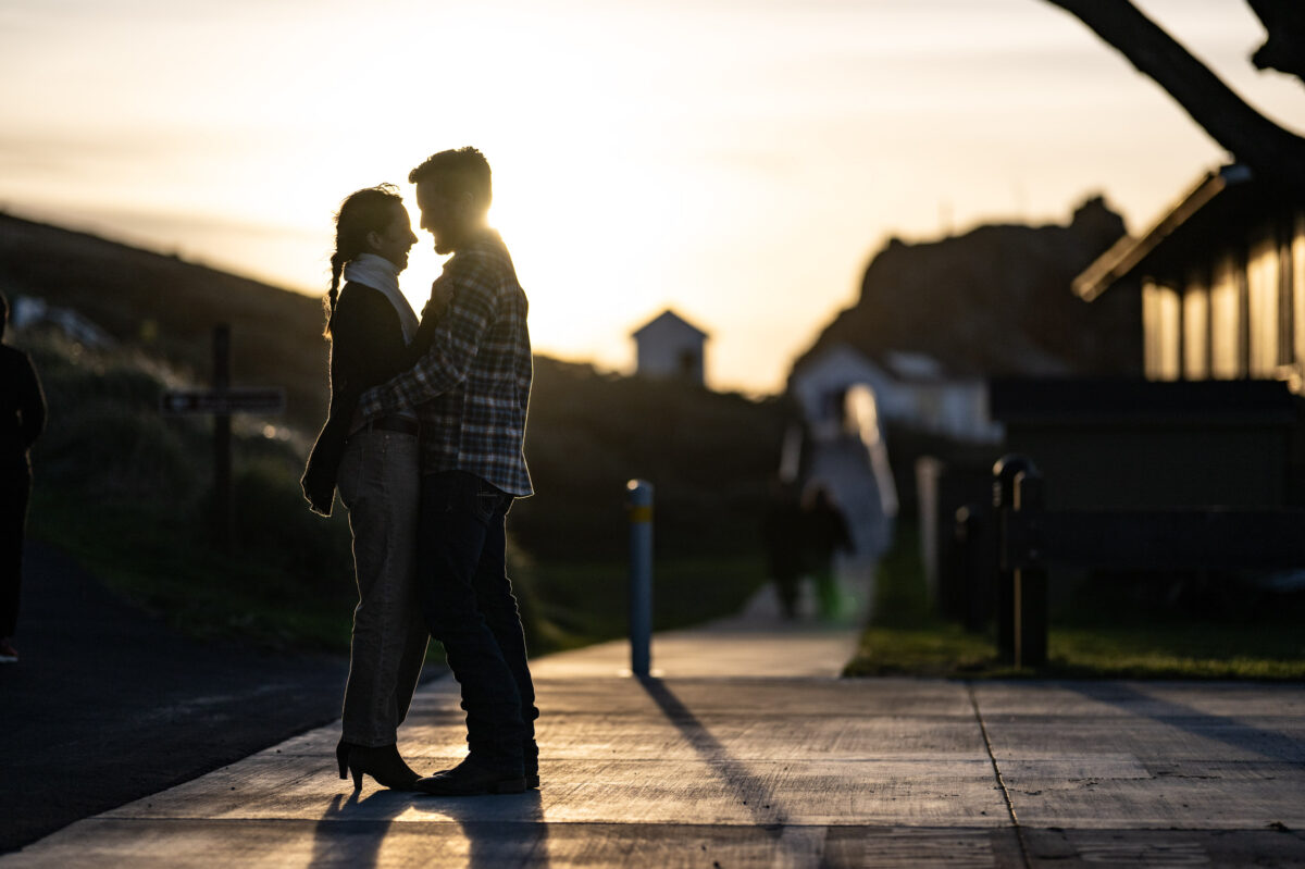 A couple stands close, facing each other, on a pathway at sunset. They are silhouetted against the glowing sky, with blurred structures and people in the background.