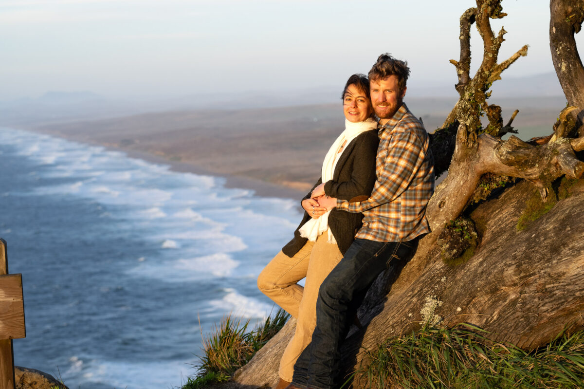 A couple leans against a tree while smiling, with an ocean and coastline visible in the background.