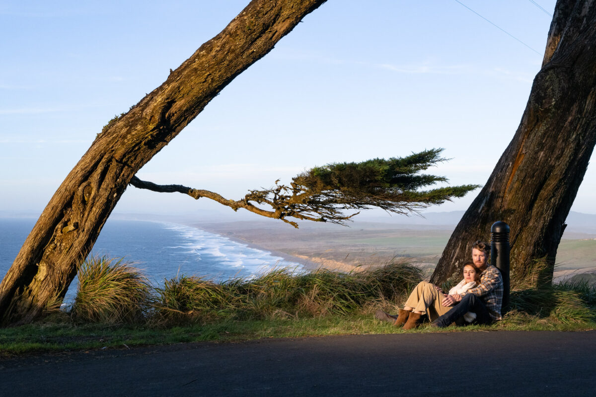 A person sits under a leaning tree, holding a baby, with an ocean and coastline in the background during daylight.