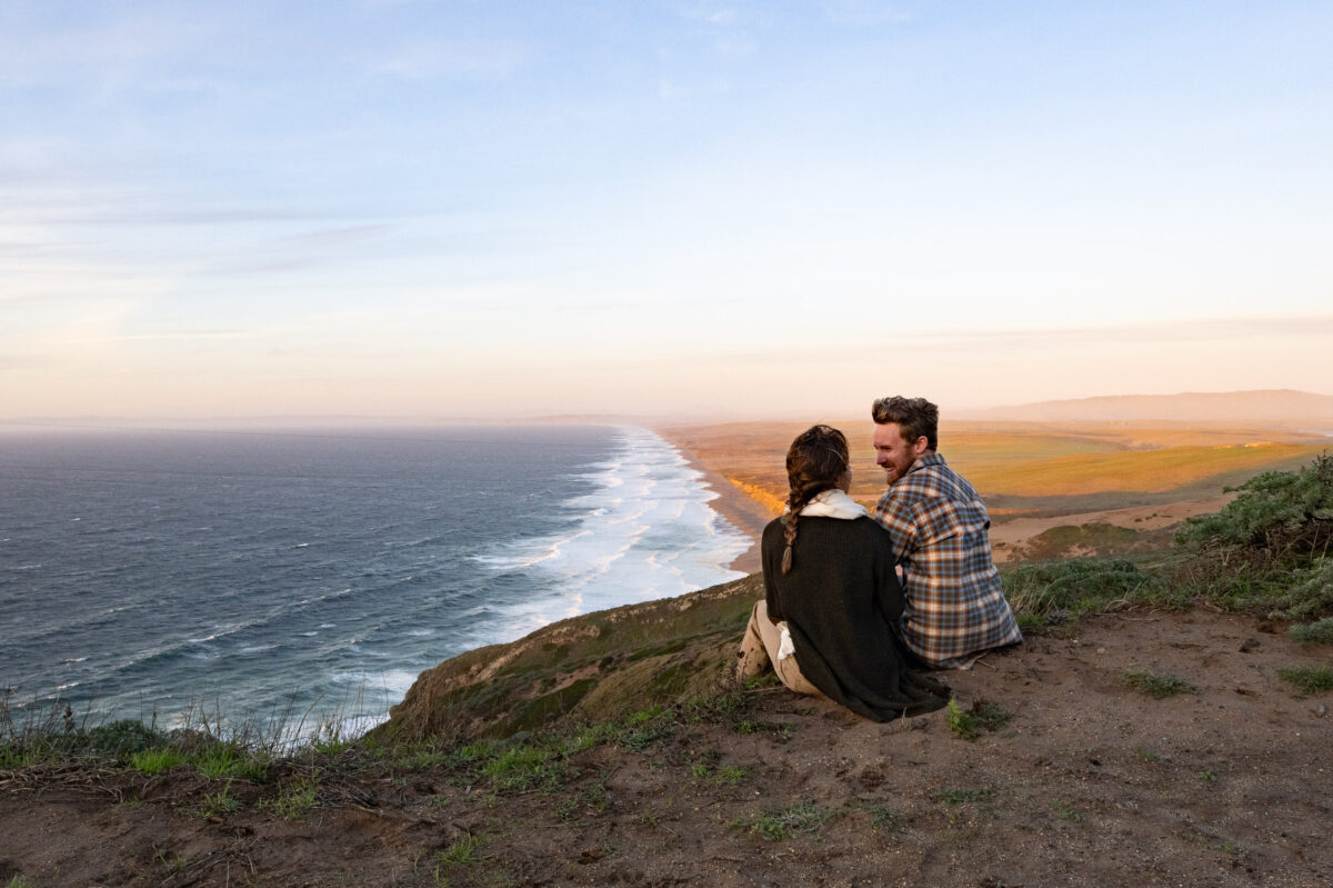 A couple sits on a grassy hilltop overlooking a coastline with waves crashing against the shore and a vast expanse of ocean under a clear sky during sunset.