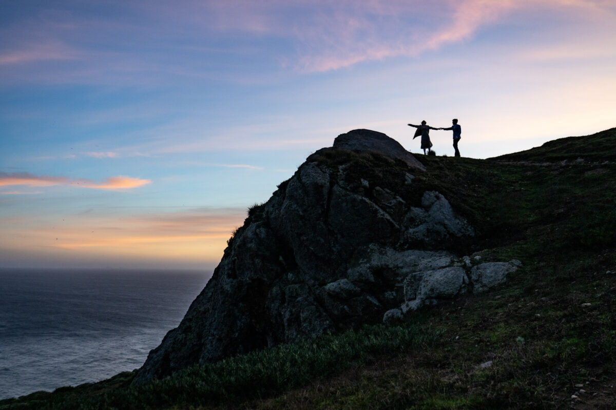 Two people stand on a rocky hill overlooking the ocean at sunset, with one person holding the other by the arm and extending their other arm outward.