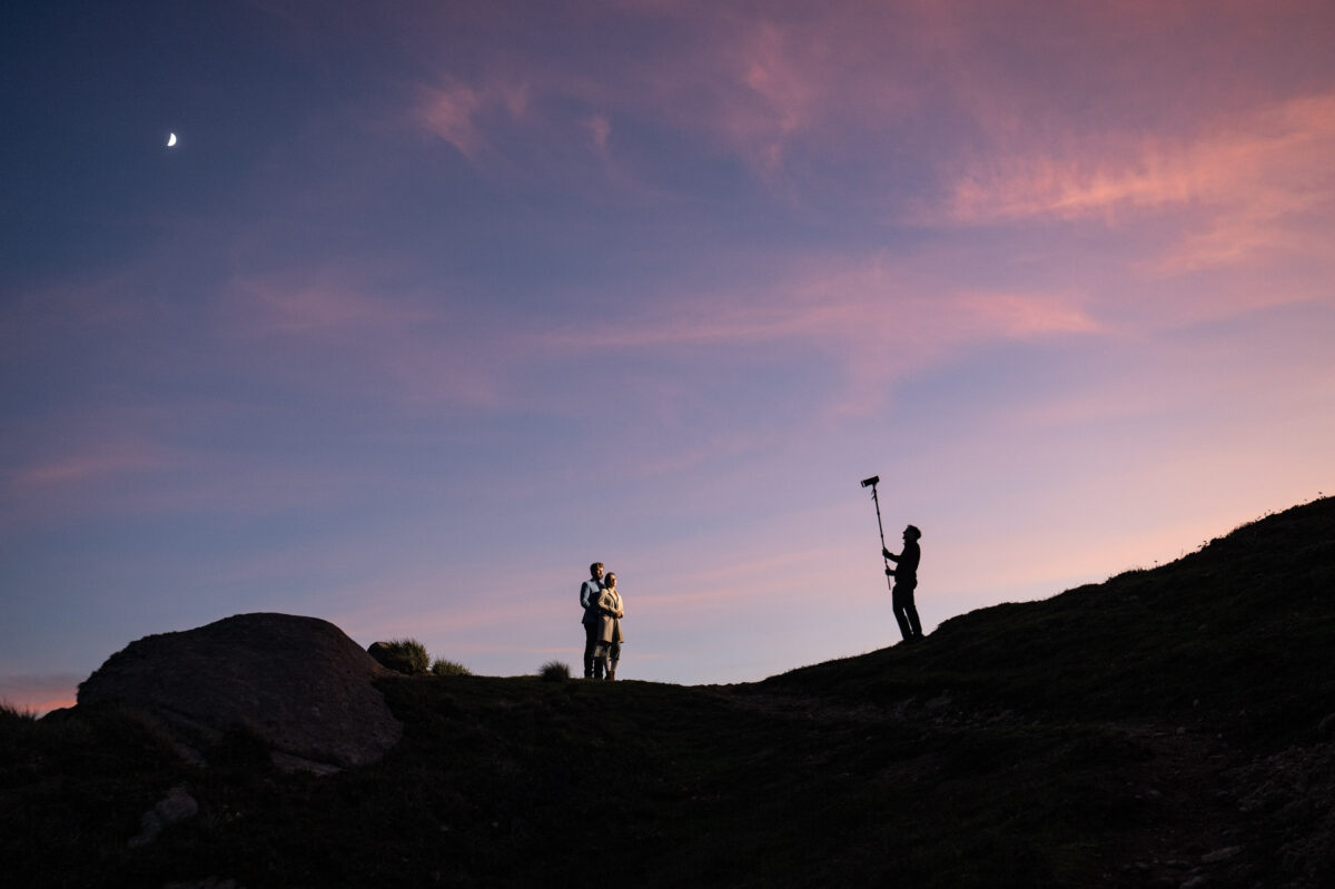 Silhouetted figures stand on a hill at sunset, one person holding a boom microphone towards a couple. A crescent moon is visible in the sky.