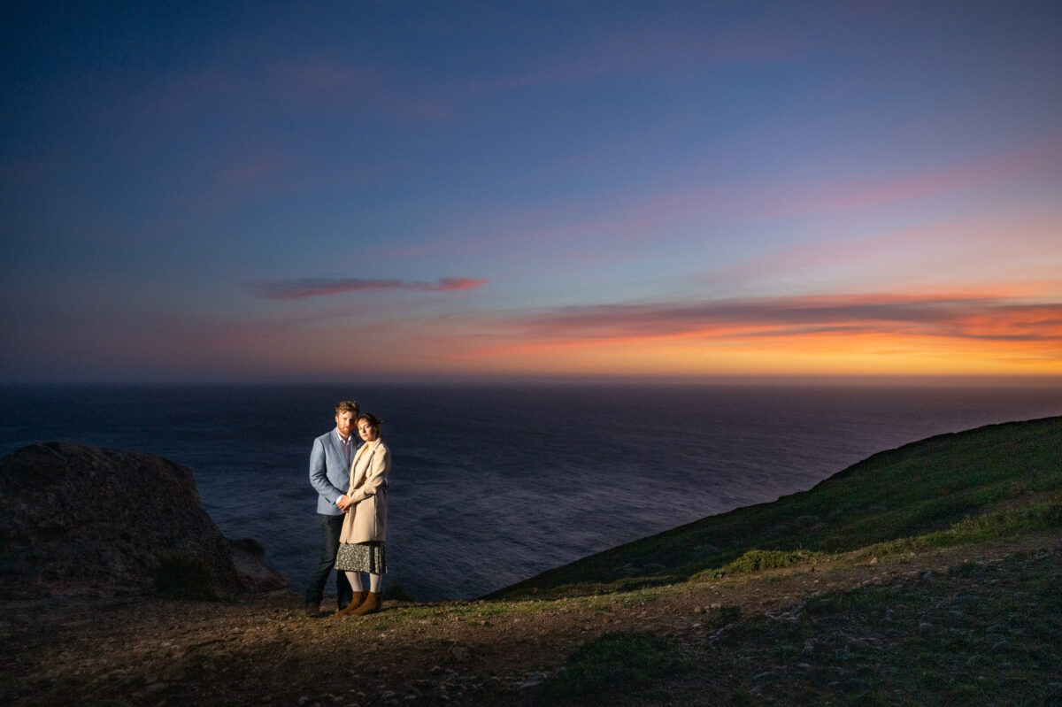 A couple stands together on a cliffside overlooking the ocean at sunset, with vibrant colors in the sky and a serene, vast seascape in the background.