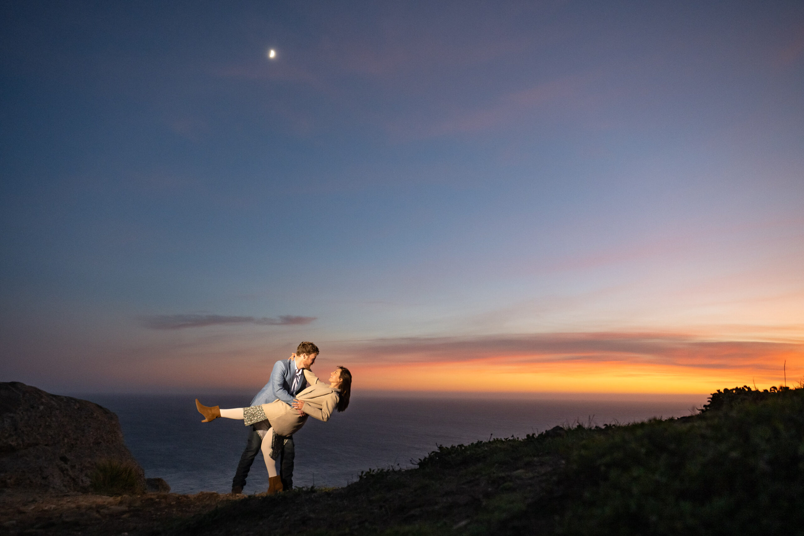 A couple poses at sunset on a cliffside, with the man lifting the woman and the moon visible in the sky above them.